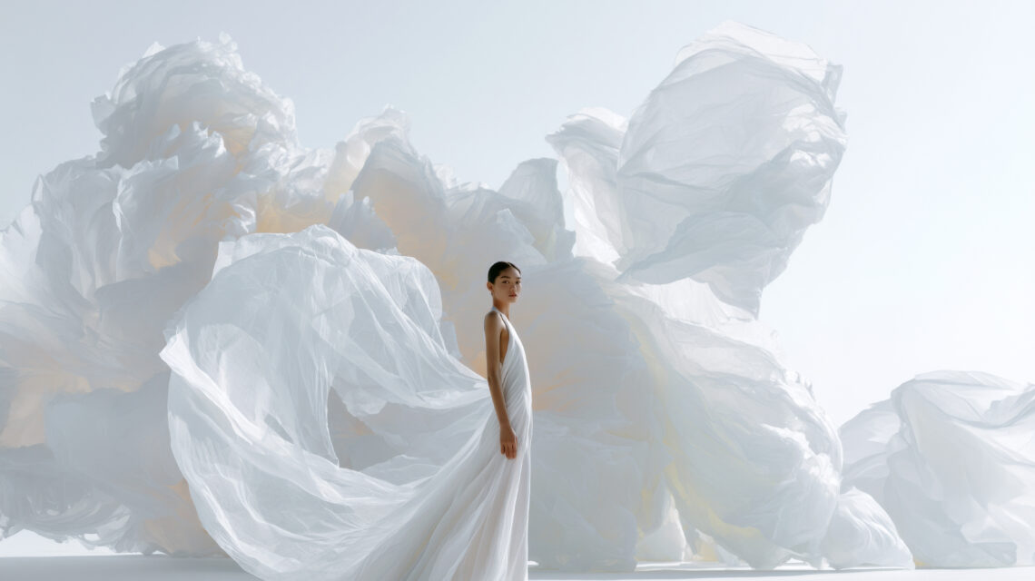 A young white-skinned woman with an elegant serene expression, full body shot from head to toe, wearing a minimal floor-length ivory white gown, standing alone on a white minimal surface, enormous sheets of ultra-thin translucent paper flying dramatically behind her caught by invisible wind, delicate fiber texture visible, paper extending beyond the frame toward a pale sky, cool white and pale ivory tones, vast negative space above, ethereal and monumental atmosphere, luxury fashion editorial photography, low angle wide shot, cinematic depth of field, soft natural lighting, ultra high resolution, photorealistic, portrait format hd --chaos 5 --ar 4:5 --raw --profile hxg5zlo --v 8.1 Job ID: 479f2762-7d1c-4968-bbca-9df200e57329